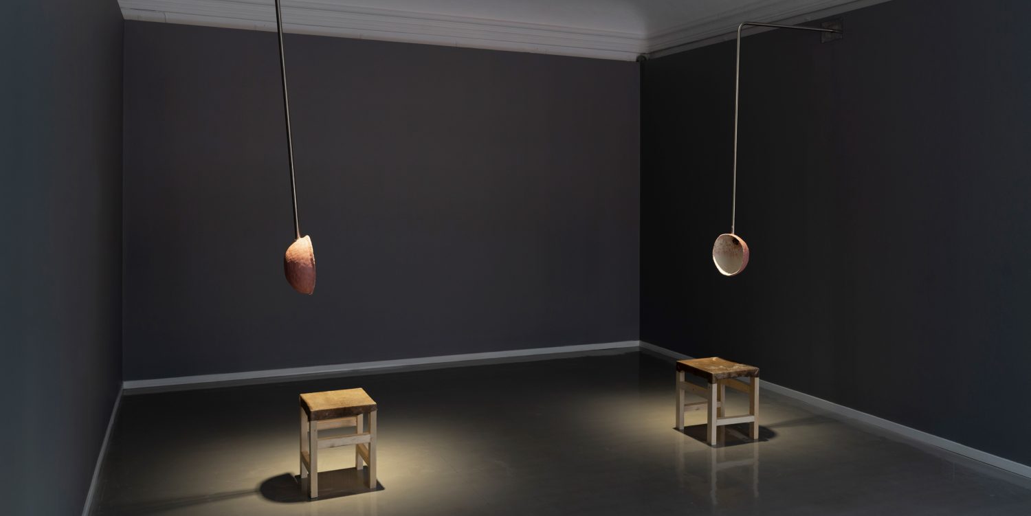 In a grey gallery room, two stools are placed on the floor. Two poles extend from the walls, each supporting a circular bowl-like object. These bowls are positioned so that a person sitting on a stool can either speak into or listen through them.