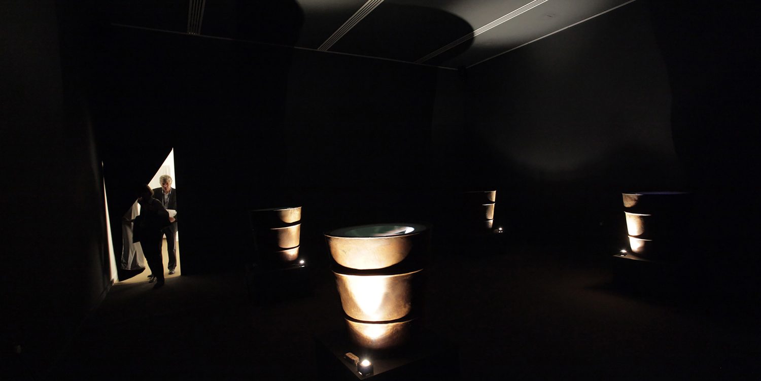 Two people enter a darkened room through a curtain, where they find four illuminated sculptures that are identical.