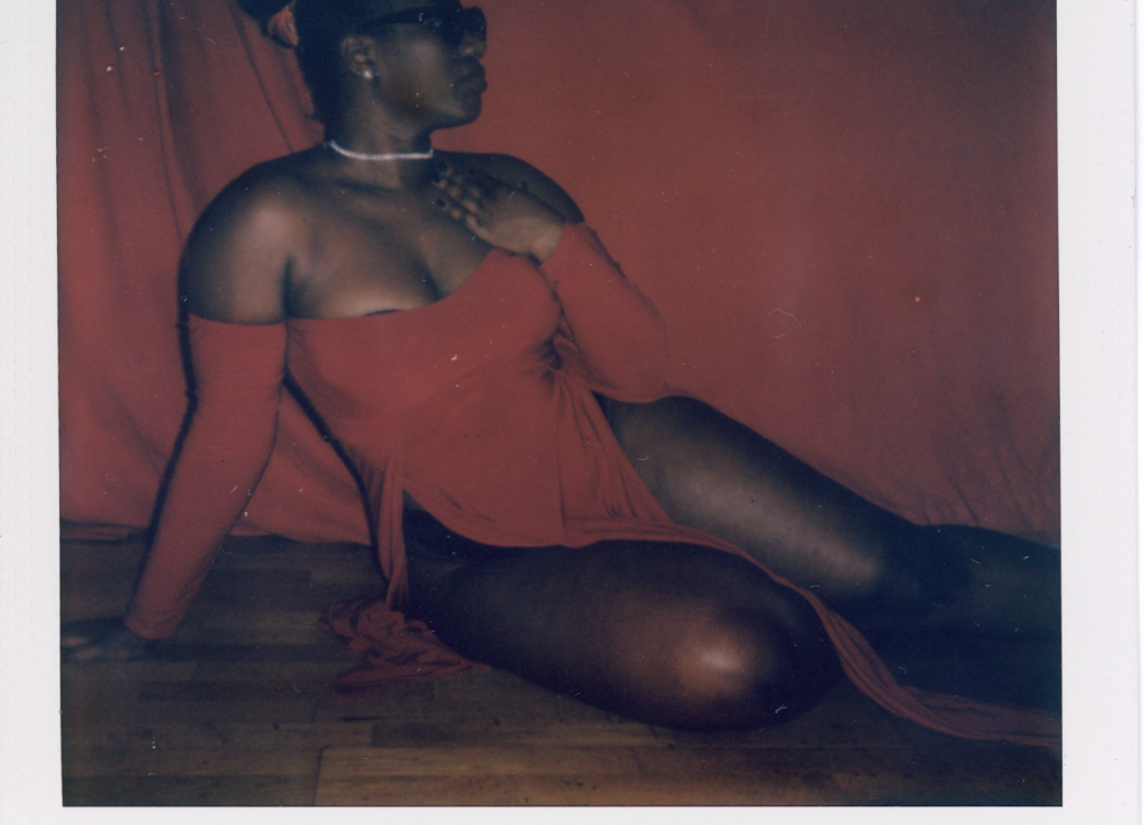 A polaroid of a woman posing on the floor in front of a red curtain. She is wearing black sunglasses and red dress.