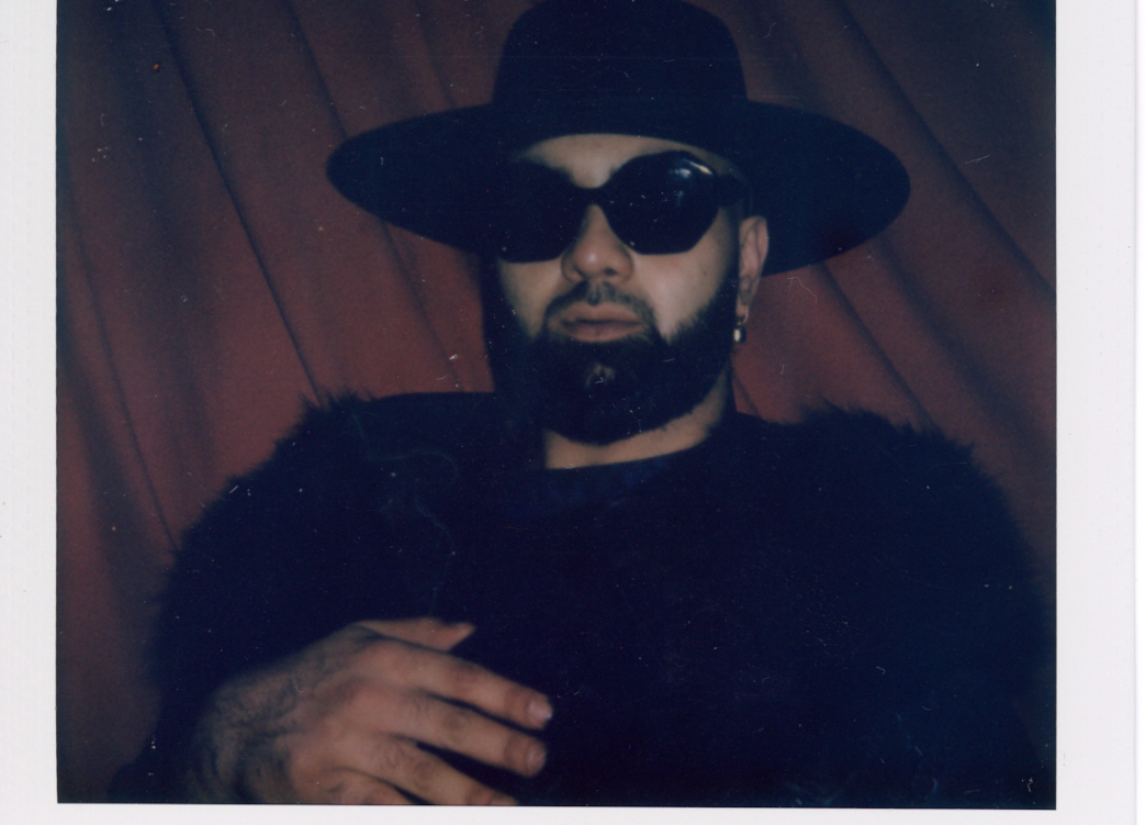 A polaroid of a man with a beard infront of a red curtain wearing a black fedora hat, black sunglasses and a black fluffy jacket.