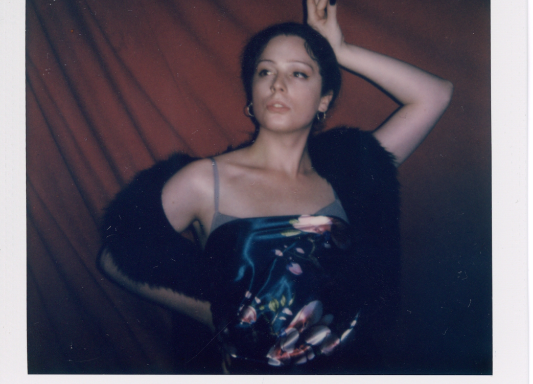 A polaroid of a woman posing with her hand above her head in front of a red curtain wearing a black fluffy scarf and a satin scarf as a top.