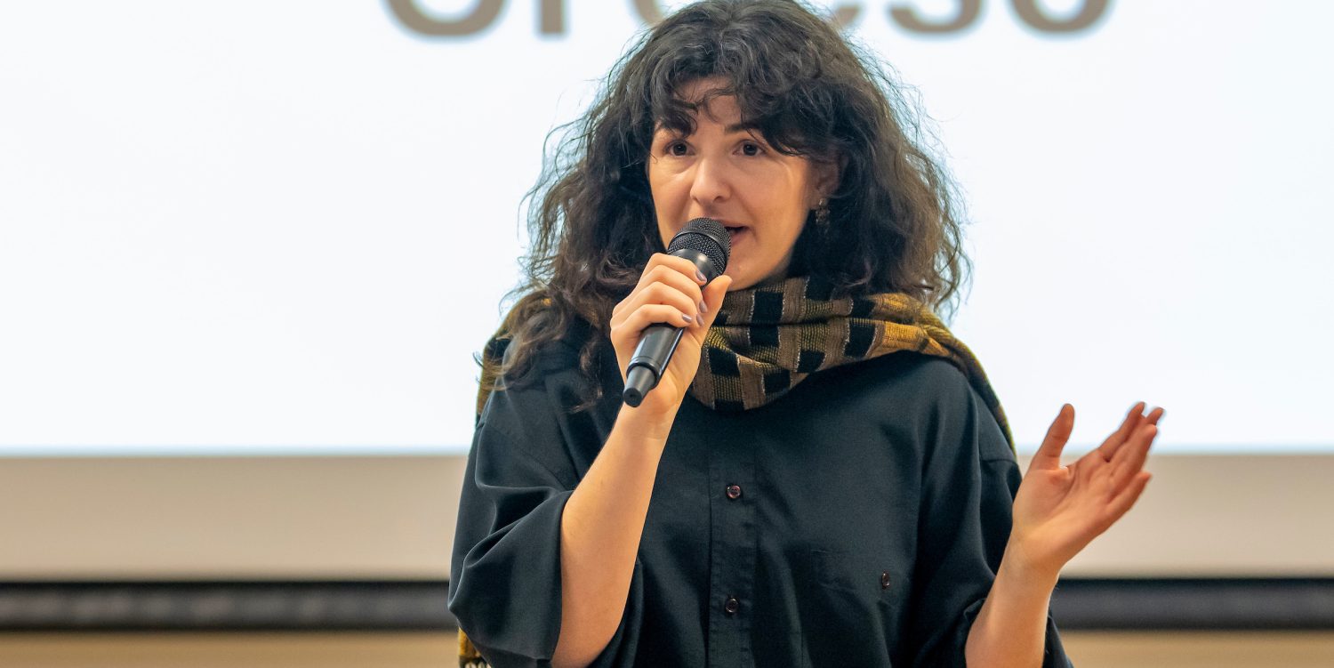 A woman standing in front of a screen that reads 'Croeso'. She is talking in to a microphone. She has long brown wavy hair and is wearing a black shirt and a scarf.