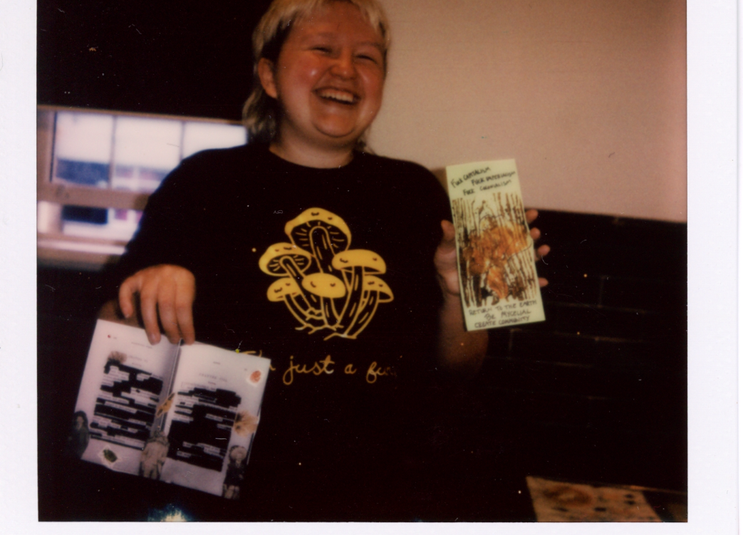 A polaroid of a woman holding up fungi publications. She is smiling and wearing a black t-shirt with a yellow mushroom graphic on it.