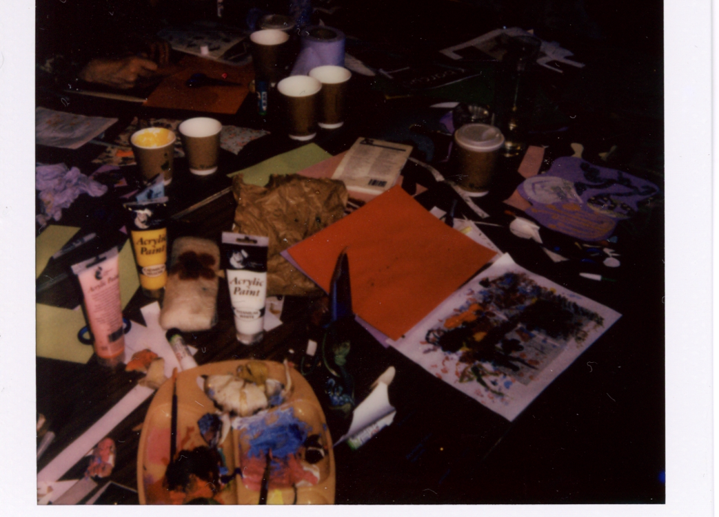 A polaroid of a table with paints, cups and other materials on top.