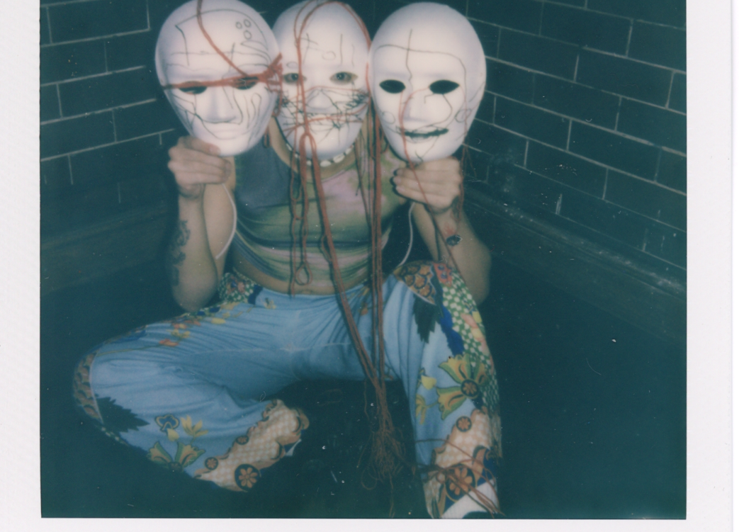 A polaroid of a person sat on the floor wearing a white mask. They're also holding up a white mask in each hand, to each side of their head.